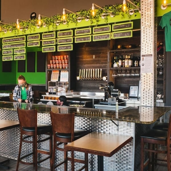 Two customers at the bar of Ineffable Brewing Co., with beer taps and brewing tanks visible.