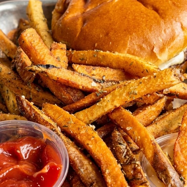A close-up of a burger and seasoned fries in a metal tray with ketchup and a creamy sauce.