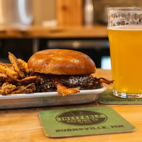 A burger with bacon and fries on a metal tray, next to a glass of beer on a wooden table.