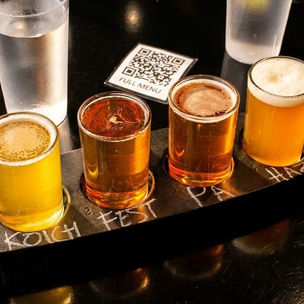 Four beer samples on a dark paddle, with a QR code menu and water glasses.