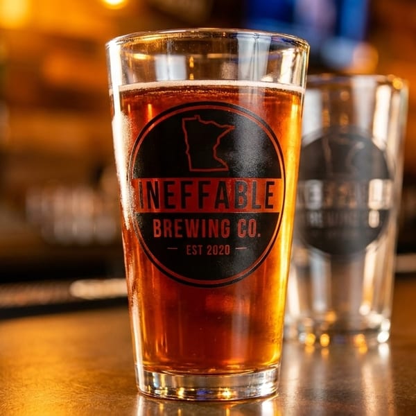 A pint glass of amber beer with an "Ineffable Brewing Co." logo sits on a bar.