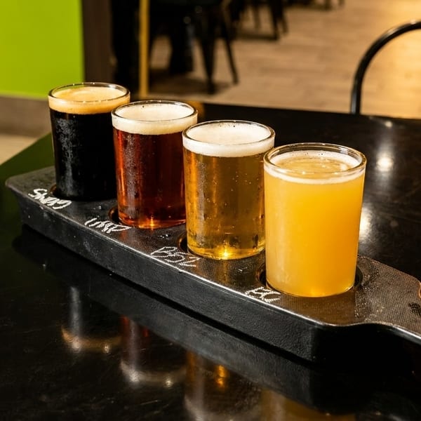 Four beer samples, ranging from dark to light, on a black paddle on a dark table.