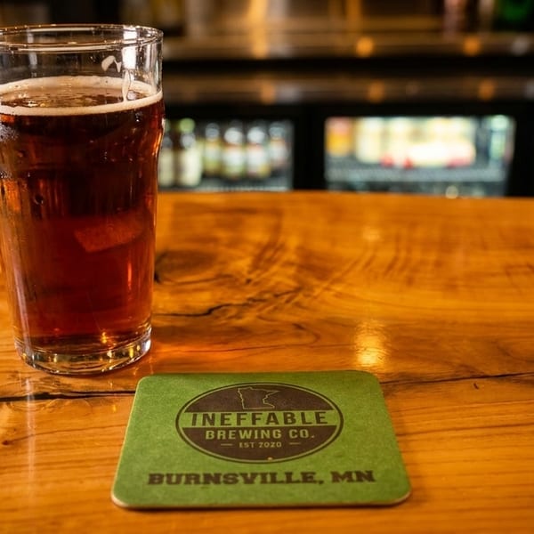 A pint of amber beer with a foamy head sits on a wooden bar next to a green coaster.